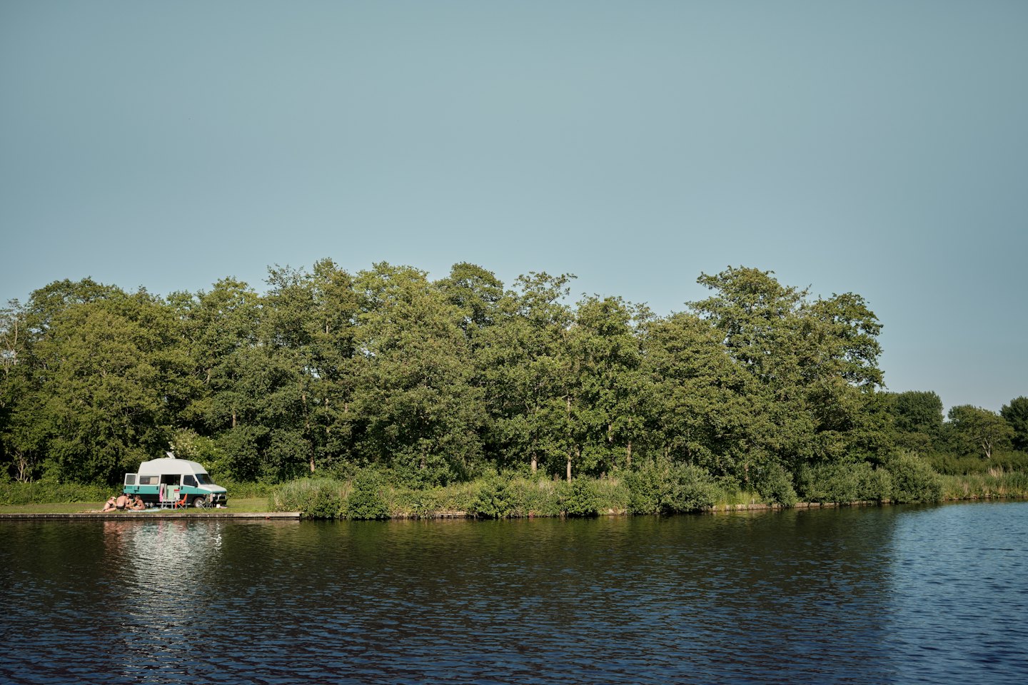 People relax by a teal and white camper van near water.