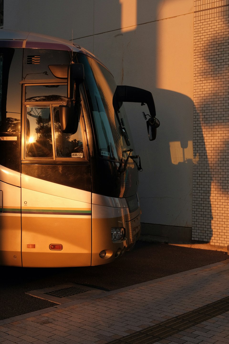 Beige and grey coach bus parked near a brick wall at sunset.